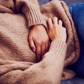 Junge Frau hat hat Unterleibskrämpfe, während sie auf der Couch liegt. Sie trägt einen hellbraunen Wollpullover und blaue Jeans, ihr Kopf ist nicht zu sehen.